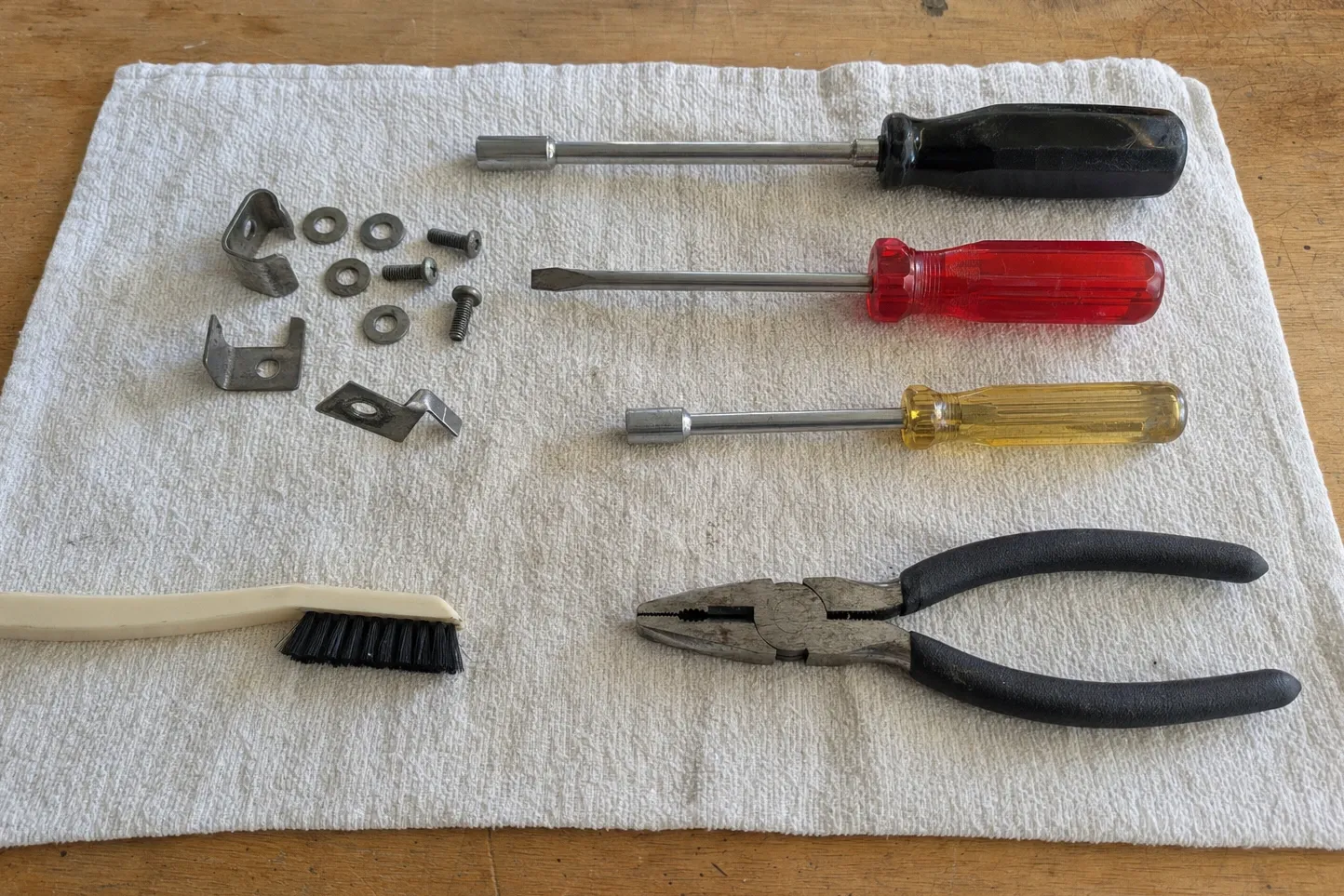 Black nut driver, red flat driver, yellow nut driver and combination pliers laid in a column on a ribbed white towel, with two small angle brackets, a Z-shaped offset clip, washers, machine screws and a cream bristle brush to their left