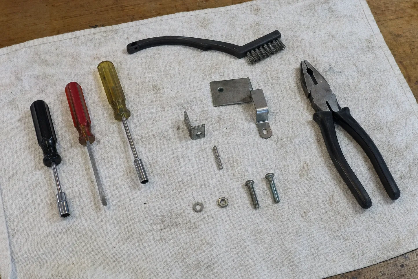 Black nut driver, red flat driver and yellow nut driver in a row on a stained work cloth, a black wire brush above them, and an angle bracket, a rectangular mounting plate, a formed clip, a pin, a washer, a nut and two machine screws in the middle with combination pliers at the right