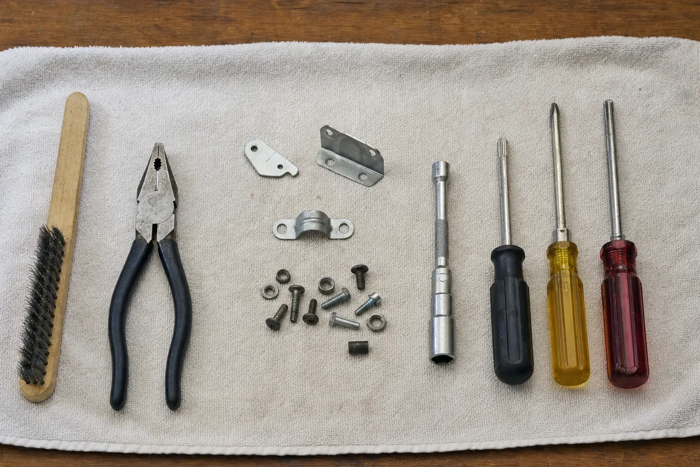 Wooden-handled wire brush and combination pliers beside a perforated link plate, a rectangular strike plate, a saddle clamp and a scattering of screws, nuts, washers and a bushing, with a telescoping socket driver, a Torx driver and two more drivers to the right on a grey towel