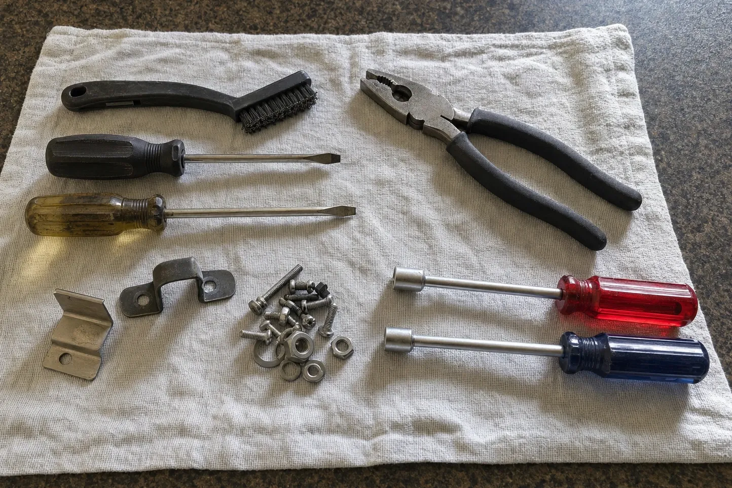 Black wire brush and two flat drivers on a cloth over a dark speckled worktop, an angle bracket and a saddle clamp with a pile of bolts, nuts and washers below them, combination pliers and two nut drivers to the right